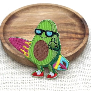 May include: An embroidered patch featuring a green avocado character wearing black sunglasses. The avocado holds a pink and yellow surfboard under one arm, has a brown pit in the center, and wears red and white sneakers on its small feet.