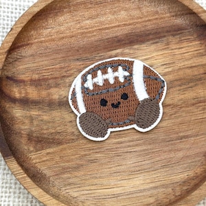 Op de afbeelding: Een geborduurde patch in de vorm van een bruine American football met witte veters en strepen. De football heeft een eenvoudig zwart geborduurd gezicht met twee kleine ronde ogen en een gebogen mond, omlijnd met wit garen.