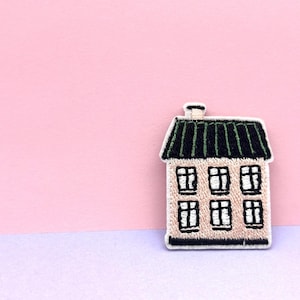 Puede incluir: Parche bordado de una casa con tejado verde y seis ventanas. La casa es beige con contornos y detalles negros. El parche está sobre un fondo rosa y morado.