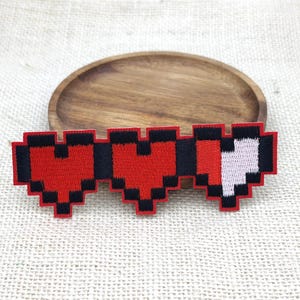 Può includere: Una toppa ricamata raffigurante tre cuori pixelati disposti in fila orizzontale. I primi due cuori sono rosso pieno con contorni neri, mentre il terzo cuore è parzialmente rosso e bianco, il tutto rifinito con un contorno nero.