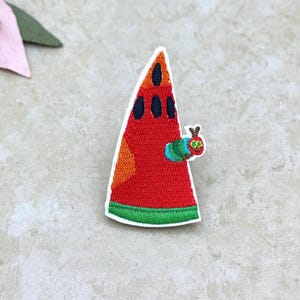 May include: A triangular embroidered patch shaped like a slice of watermelon with a red center, black seeds, an orange side, and a green rind. A small, multi-colored caterpillar with a red head and green body is attached to the side of the slice.