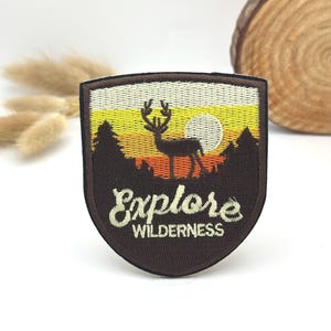 May include: Embroidered patch with a shield shape, featuring a deer silhouette against a sunset backdrop. The patch includes the text "Explore Wilderness" in a stylized font, with a brown border and a cream-coloured background.