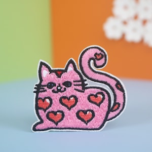Puede incluir: Parche de gato bordado de color rosa con contorno negro y corazones rojos. El gato está sentado con la cola enroscada alrededor de su cuerpo.