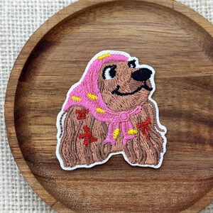 May include: An embroidered patch featuring a brown dog wearing a pink headscarf with yellow accents. The dog has a black nose and eyes, and the fur is depicted with brown thread. The patch has a white border and is shaped like the dog's head and chest.