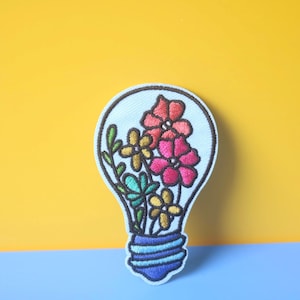 Puede incluir: Parche bordado con forma de bombilla, lleno de flores coloridas. Las flores incluyen flores rojas, amarillas y turquesas con hojas verdes. El parche tiene un fondo blanco y una base azul, sobre un fondo amarillo y azul.