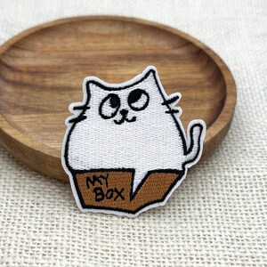 Puede incluir: Parche bordado con un gato blanco con contornos negros, sentado en una caja marrón con la inscripción "MY BOX". El parche está sobre un plato de madera.
