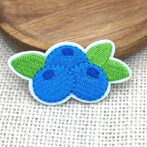 May include: Embroidered patch featuring a cluster of three blueberries in shades of blue, with green leaves. The patch has a white border and is likely for clothing or accessories.