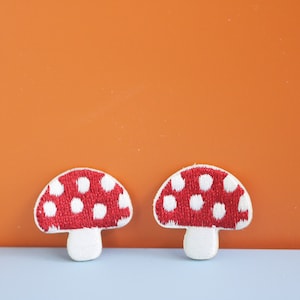 May include: Two embroidered patches shaped like mushrooms. Each patch features a red cap with white polka dots and a white stem. The embroidery is dense and textured, creating a clear outline for each mushroom shape against the background.