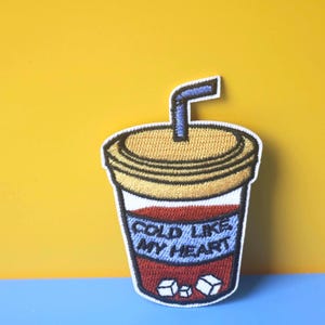 May include: A colorful embroidered patch of a cold drink with a straw and ice cubes. The text on the patch reads "Cold Like My Heart".