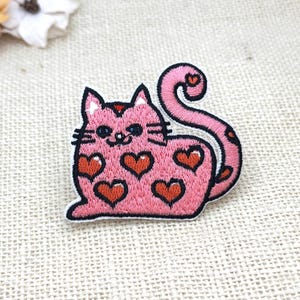 May include: An embroidered patch featuring a pink cat with a long, curled tail. The cat has dark eyes, whiskers, and a body covered in multiple red heart shapes with black outlines. The entire design is bordered by a thick black stitched edge.