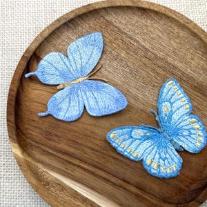 May include: Two embroidered butterfly patches resting on a round wooden tray. The top butterfly is light blue with textured stitching. The bottom butterfly is a brighter blue with gold-colored accents along the wing edges and intricate thread details.