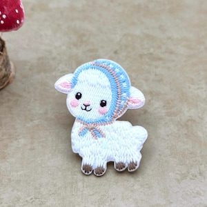 Pode incluir: Remendo bordado de um cordeiro branco usando um capuz azul com detalhes em rosa. O cordeiro tem uma expressão doce com bochechas rosadas e cascos marrons. O remendo é provavelmente para roupas ou acessórios.