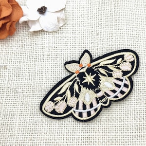 May include: Embroidered moth patch with a black border, showcasing cream, peach, and gold detailing. The moth has a star design in the centre and is set against a neutral background with a rose and a white flower.