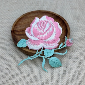 Puede incluir: Un parche bordado que muestra una rosa en flor con pétalos superpuestos de color rosa y blanco. La flor está unida a un tallo con hojas de color verde claro y un pequeño capullo de rosa sin abrir. El bordado utiliza puntadas densas para crear textura.