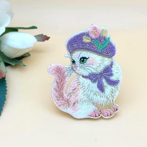 Op de afbeelding: Geborduurde patch van een wit en roze katje met een paarse hoed en strik. De kat heeft groene ogen en een roze staart. De patch staat op een lichtbeige achtergrond. Een witte roos en groene bladeren staan in de linkerbovenhoek.