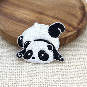 May include: An embroidered patch featuring a stylized panda bear in a prone position. The panda is rendered in black and white thread, showcasing a round head with dark patches around the eyes, small ears, and a rounded body with black limbs.