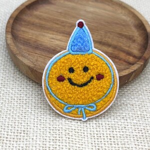 Puede incluir: Un parche bordado circular que presenta una cara sonriente amarilla con una sonrisa cosida en negro, dos mejillas rojas pequeñas y un gorro de fiesta azul en la parte superior. El parche tiene un borde azul claro y un lazo azul en la base.