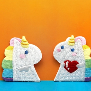 May include: Two white embroidered unicorn patches. One unicorn is holding a red heart. Both unicorns have rainbow manes.