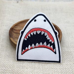 May include: A white, embroidered shark patch with a black outline. The shark's mouth is open, revealing rows of sharp, white teeth and a red-lined mouth. The patch is on a wooden plate.