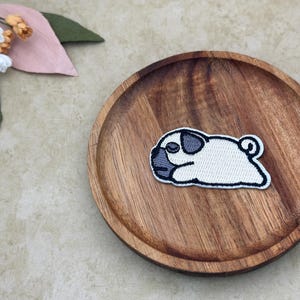 以下が含まれることがあります： 木製のプレートの上に横たわる、黒い縁取りのある白とグレーのパグのパッチ。 パッチは詳細なデザインで刺繍されています。 背景には装飾的な花と葉があります。