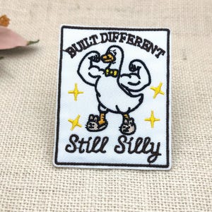 May include: A rectangular embroidered patch with a white background and black border. It features a muscular white duck wearing a yellow bowtie and slippers, surrounded by yellow stars. The text reads BUILT DIFFERENT and Still Silly.