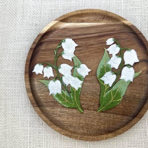 Può includere: Due toppe ricamate di mughetto, con fiori bianchi e foglie verdi, sono esposte su un piatto rotondo di legno. Le toppe sono dettagliate con cuciture delicate e accenti dorati.