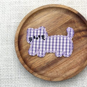 May include: A fabric cat applique with a purple and white gingham pattern, outlined in purple, sits on a round wooden plate. The cat has black eyes and whiskers. The plate is on a white textured surface.