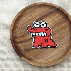 May include: A red embroidered alligator patch with a white border, featuring angry eyes and sharp teeth. The patch is placed on a round, wooden plate with a natural wood grain finish. The background is a textured, off-white fabric.