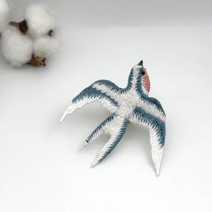 Może przedstawiać: Haftowana naszywka z jaskółką z białymi, niebieskimi i różowymi detalami. Ptak ma rozłożone skrzydła i jest wykonany ze szczegółowymi szwami. Naszywka znajduje się na białej powierzchni, z pąkami bawełny w tle.