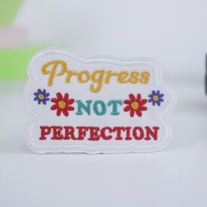 May include: White embroidered patch with the text "Progress not perfection" in yellow, red, and blue lettering. The patch is surrounded by a white border and features two red and yellow flowers with blue centres on either side of the text.