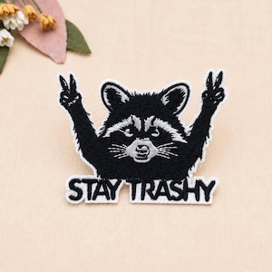 Op de afbeelding: Een geborduurde patch met een zwart-witte wasbeer die zijn armen omhoog houdt met vredestekens. De wasbeer heeft een gedetailleerd gezicht met snorharen en een witte snuit. Onder de wasbeer is de tekst STAY TRASHY geborduurd in dikke zwarte letters.