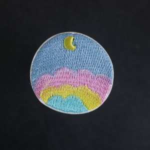 May include: A round embroidered patch with a blue sky, pink and yellow clouds, and a yellow crescent moon.