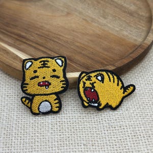Op de afbeelding: Twee geborduurde patches met cartoon tijgerontwerpen. De ene patch toont een staande tijger met een kleine mond en witte buik, terwijl de andere een hurkende tijger toont met een wijd open mond die een rode binnenkant en tanden laat zien.