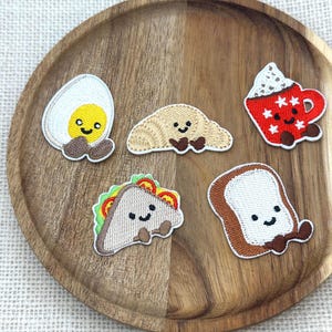 May include: A collection of embroidered patches featuring breakfast foods. Includes a fried egg, croissant, coffee cup, sandwich, and toast. Each patch has a smiling face and is set on a wooden tray.