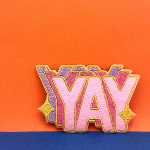 Peut inclure: Un patch "YAY" rose et violet avec un contour doré. Le patch est sur un fond orange.