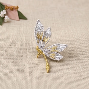 May include: An embroidered dragonfly patch featuring metallic silver and gold thread. The insect design displays detailed wing patterns with silver outlines and gold centers, complemented by a slender gold body and a pointed tail section.