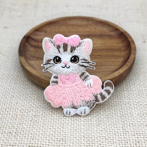 Peut inclure: Un écusson brodé représentant un chaton tigré brun et blanc aux grands yeux sombres, portant une robe rose clair et un nœud rose assorti sur la tête. Le chaton a une queue rayée et est représenté en position assise avec des pattes blanches.