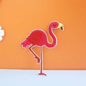 Puede incluir: Un parche bordado de flamenco rosa con pico amarillo y ojos blancos. El flamenco está de pie sobre una pierna con la otra doblada.