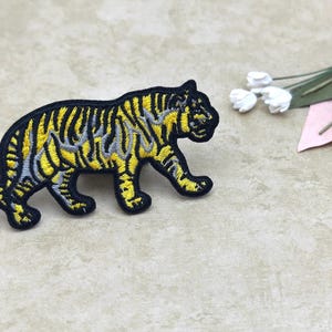 May include: Embroidered tiger patch in black, yellow, and gray. The tiger is depicted in a walking pose with detailed stripes. The patch is likely for clothing or accessories.