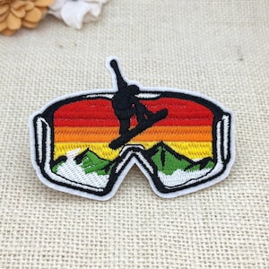 Può includere: Una toppa ricamata a forma di maschera da sci che presenta la sagoma nera di uno snowboarder che esegue un trick su uno sfondo al tramonto a strisce orizzontali rosse, arancioni e gialle, con montagne verdi e neve bianca alla base.