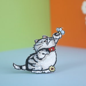Op de afbeelding: Een geborduurde strijk-op patch van een witte en grijze kat met zwarte ogen en een rode halsband, die omhoog reikt om een blauwe vlinder te vangen. De kat zit op een witte bloem.
