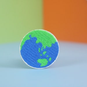 以下が含まれることがあります： 青い海と緑の大陸を持つ地球を描いた丸い刺繍パッチ。パッチは白で、白い縁取りがされています。