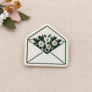 May include: An embroidered patch shaped like an open envelope with a cream-coloured base and dark green stitched borders. Inside the envelope, there is a detailed floral arrangement featuring white daisy-like flowers with yellow centres and green leaves.