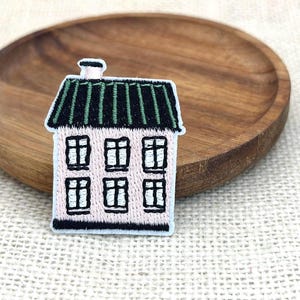 May include: Embroidered house patch with a pink facade, black windows, and a green roof. The patch has a white border and a small chimney. The patch is placed on a wooden plate.