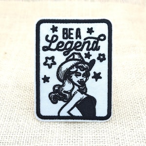 Könnte beinhalten: Ein rechteckiger bestickter Aufnäher mit weißem Hintergrund und schwarzem Rand. Das Design zeigt eine schwarze Illustration einer Person mit Cowboyhut, umgeben von kleinen Sternen, mit dem in Schwarz geschriebenen Text BE A Legend.