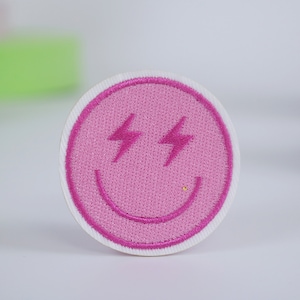 May include: Pink embroidered smiley face patch with lightning bolt eyes. The patch is circular and has a white border.