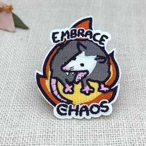 Op de afbeelding: Geborduurde patch met een cartoonbuidelrat omringd door vlammen. De buidelrat is grijs en wit met roze accenten, en de patch bevat de woorden "EMBRACE CHAOS" in witte letters. Witte rand, oranje en gele vlammen.