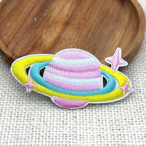 May include: Embroidered patch featuring a colorful planet design. The planet is pink and white with a yellow and blue ring. Pink star accents are on the patch. The patch is on a light beige surface with a wooden background.