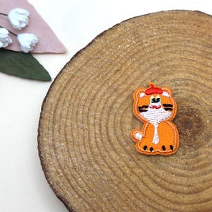 May include: An orange embroidered patch in the shape of a tiger wearing a red beret. The patch is on a wooden slice with a natural wood grain pattern. Decorative flowers are in the background.