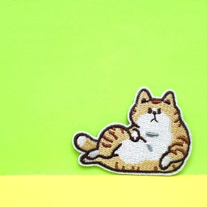 May include: A brown and white embroidered patch of a cat lying on its side with its paws tucked in. The cat has a brown nose and black eyes.